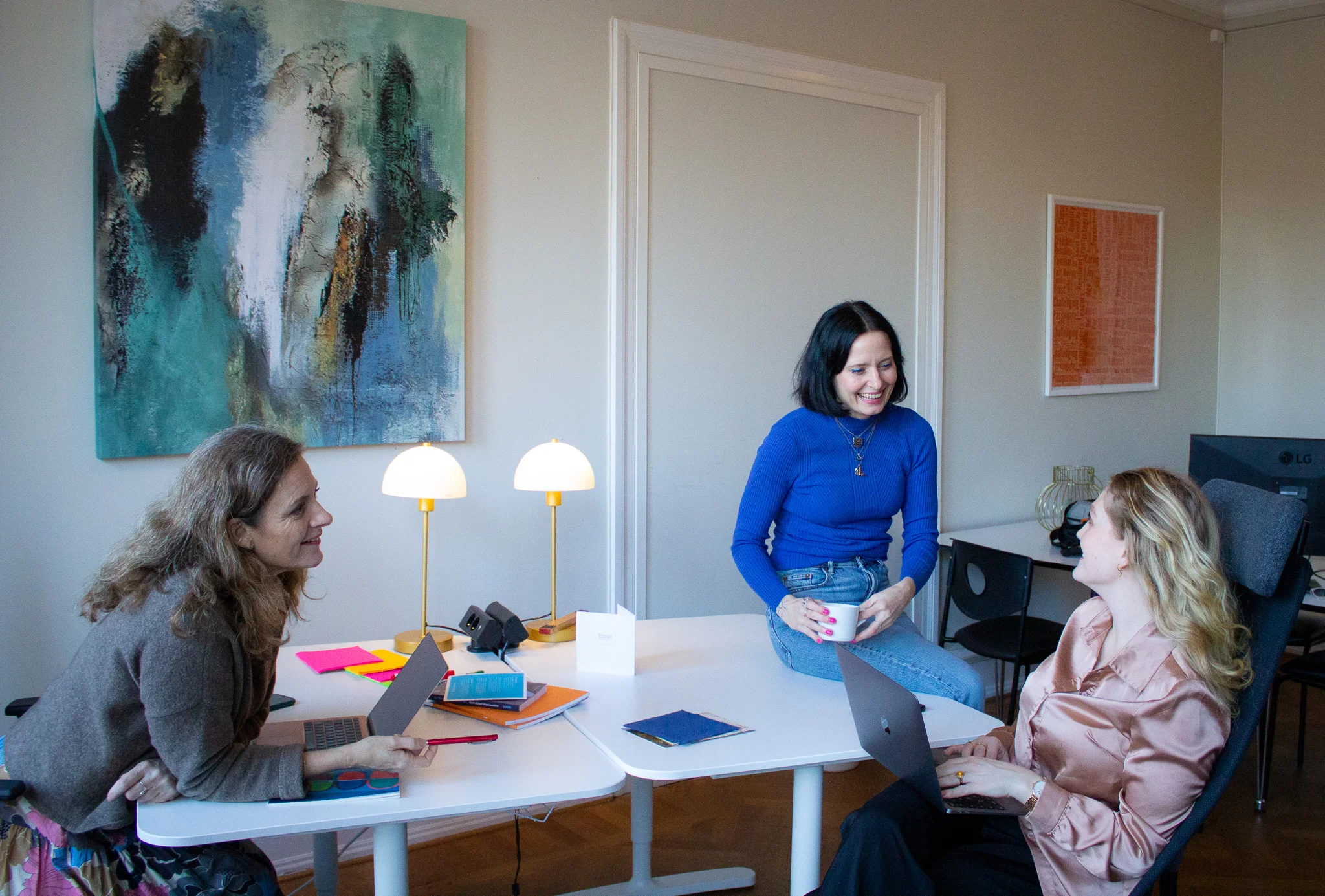 Kreativt teamarbete på eventbyrån Cronström & Co - tre kvinnor planerar strategiska event runt bord med laptops och färgglada anteckningar. Engagerad diskussion i ljust Malmö-kontor med modern inredning och konstnärliga detaljer